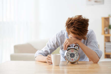 Sleepy young man with alarm clock sitting at the tableの写真素材