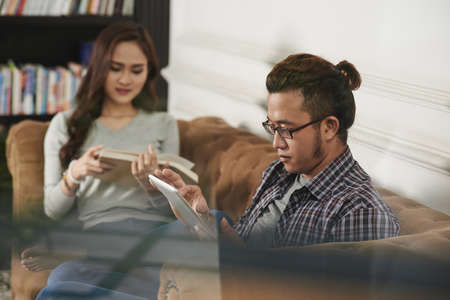 Young man reading on tablet computer while his girlfriend reading paper bookの写真素材