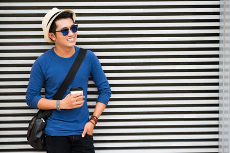 Cheerful man with crossbody bag and take-out coffee standing at the striped wallの写真素材