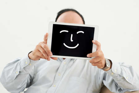 Senior man hiding his face behind digital tablet with drawn smileの写真素材