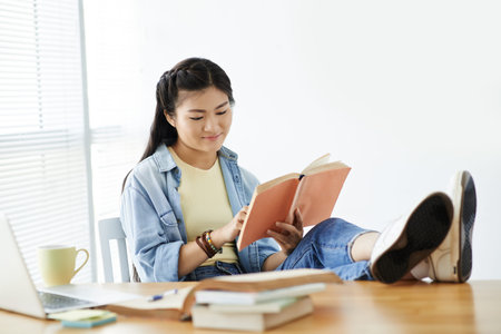 College girl sitting with legs on her table and reading a book