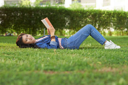 Pretty Asian student lying on grass with her favorite book and smiling at the cameraの写真素材