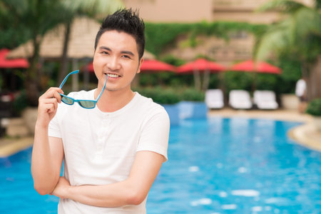 Portrait of happy Asian man with sunglasses standing at the poolの写真素材