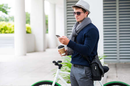 Stylish young man with paper cup of coffee using application on his smartphoneの写真素材
