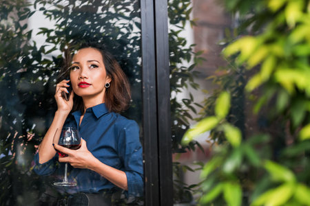 Good-looking lady drinking wine and enjoying view from the window when calling on the phoneの写真素材