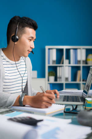 Vietnamese businessman in headset working on laptop and making notesの写真素材