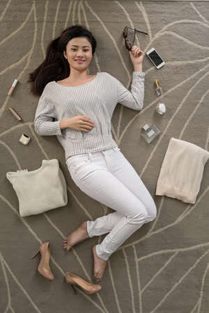 Smiling Asian woman lying on the floor with cosmetics and sweaters around herの写真素材