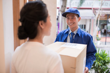Cheerful Asian postman giving cardboard box to the womanの写真素材