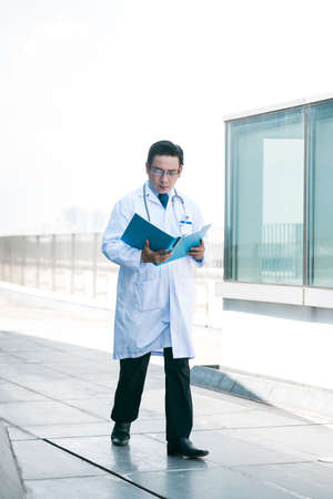 Serious medical worker walking outdoors and reading medical historyの写真素材