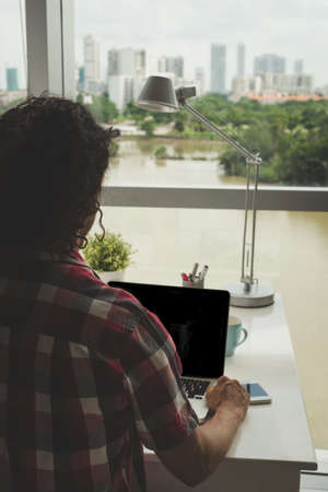 Man working on laptop in front of large window, view over the shoulderの写真素材