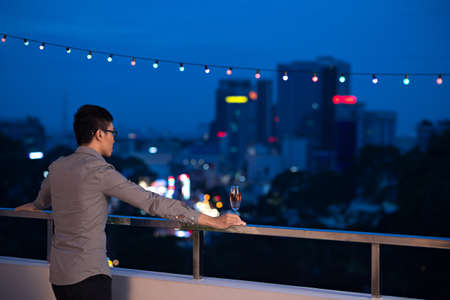 Rear view of man with a glass of champagne standing on the balcony and enjoying the view of the night cityの写真素材