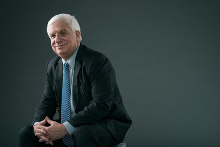Smiling senior businessman sitting and looking at the camera, isolated on grey