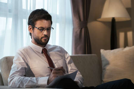 Businessman in glasses sitting on the sofa and making notesの写真素材