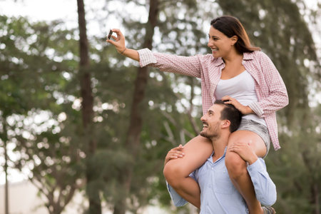 Pretty young woman sitting on the shoulders of her boyfriend and taking selfieの写真素材