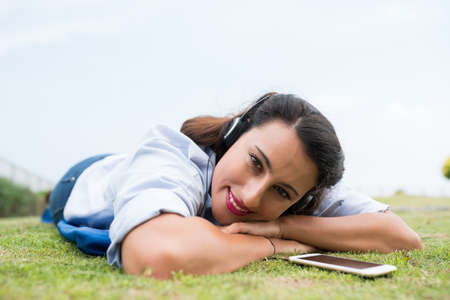 Cheerful woman in headphones lying on the grass and looking at the cameraの写真素材