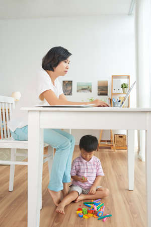 Vietnamese woman working on laptop when her son playing under the tableの写真素材