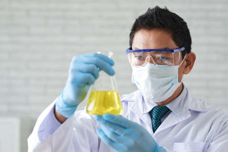 Serious Asian scientist looking at beaker with yellow liquidの写真素材