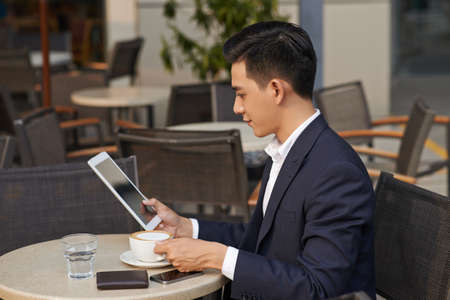 Elegant Asian man reading something on tablet computer when drinking coffee in restaurantの写真素材