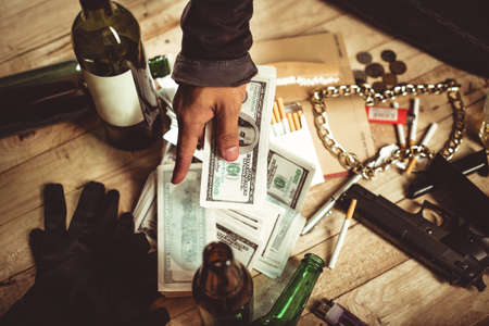 Close-up of male hand holding dollars which lie on table among gun and bottles of wineの写真素材