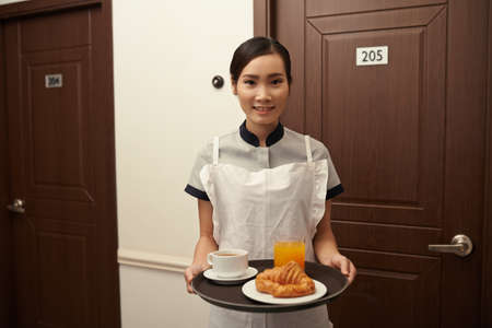 Young smiling maid in apron holding tray with breakfastの写真素材