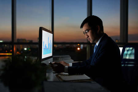 Serious Vietnamese businessman working on financial report in dark officeの写真素材
