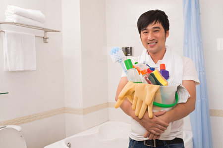 Smiling Asian guy with basket of cleaning supplies standing in bathroomの写真素材