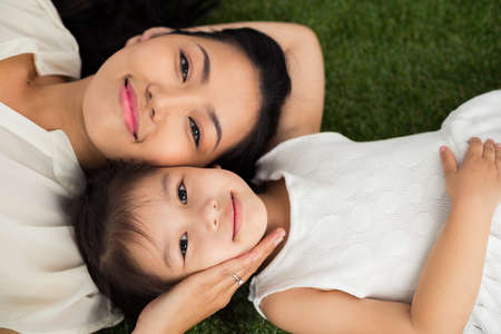 Faces of pretty Asian mother and daughter lying on grassの写真素材