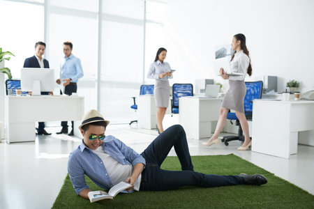 Man lying on green meadow in the center of the office and reading a bookの写真素材