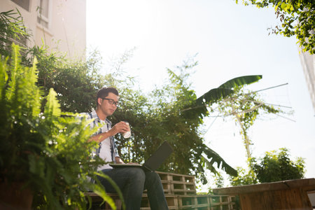 Asian man drinking take-out coffee and working on laptop outdoorsの写真素材
