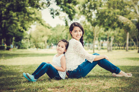 Beautiful young mother and her daughter sitting on the grass back to backの写真素材