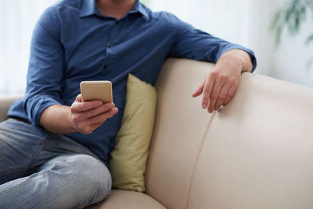 Cropped image of young man checking his phone when sitting on the sofaの写真素材