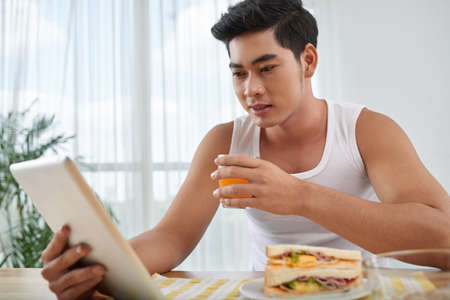 Young man drinking glass of juice and reading something on digital tabletの写真素材