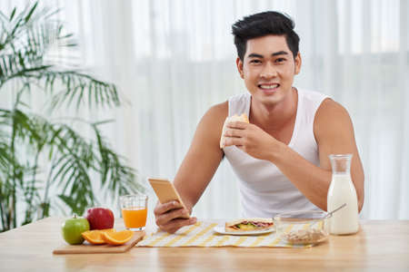 Portrait of smiling Vietnamese man with smartphone having breakfastの写真素材