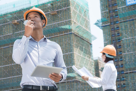 Vietnamese supervisor with digital tablet using walkie-talkie at construction site