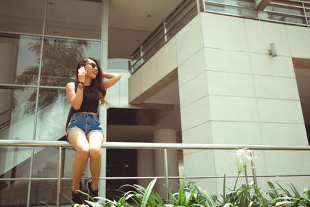 Cheerful young woman sitting on railing and talking on phone with friendの写真素材