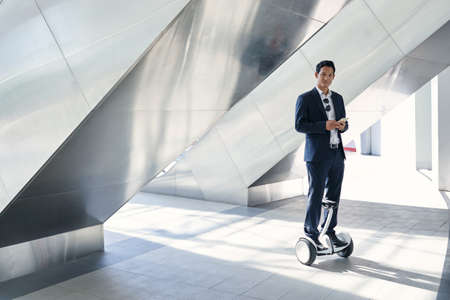 Happy businessman with cellphone standing on hoverboard in urban environmentの写真素材