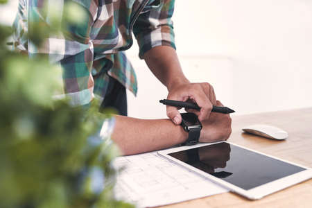 Young designer looking at his watch while wroking over blueprintの写真素材