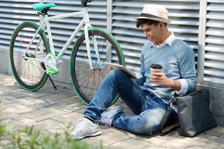 Smiling Asian guy sitting outdoors and using app on tabletの写真素材