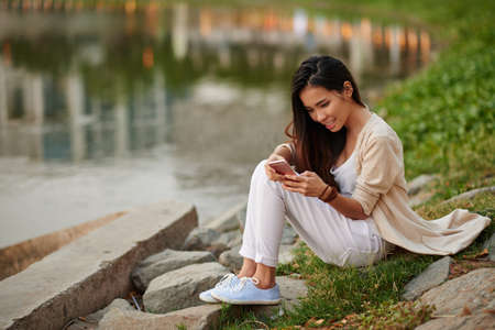 Vietnamese young woman sitting at river and using app in her phoneの写真素材