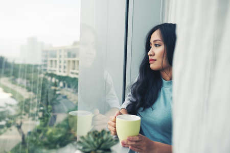 Vietnamese young woman drinking coffee and looking through windowの写真素材