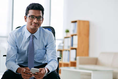 Portrait of smiling Indian business person with smartphone sitting in officeの写真素材