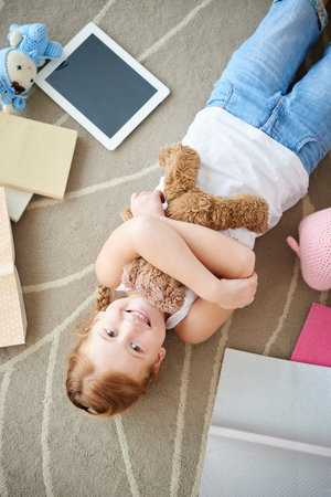 Smiling girl embracing her bear when lying on floorの写真素材