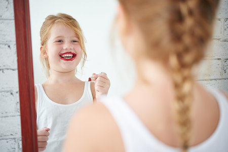 Cheerful little girl applying red lipstick in front of mirrorの写真素材