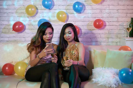 Party girls sitting on sofa and watching something on smartphoneの写真素材