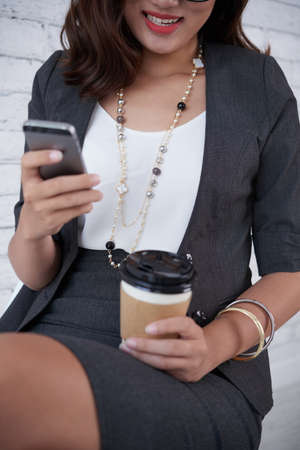 Cropped image of smiling business woman enjoying coffee when checking smartphoneの写真素材