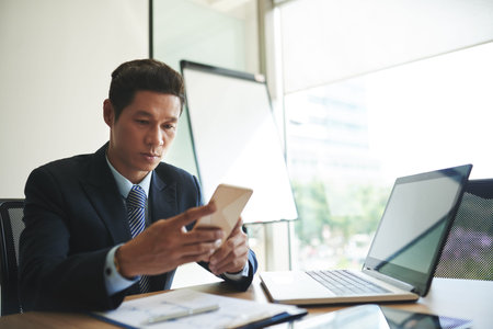 Serious Vietnamese businessman sitting at workplace and reading message on his smartphoneの写真素材