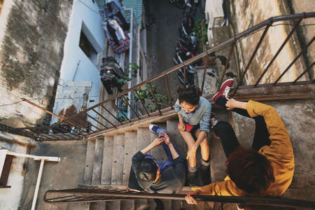 High angle view of stylish Asian teenagers hanging out in urban slums: they sitting on stairs of shabby apartment house and talking to each otherの写真素材