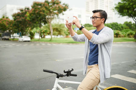 Smiling Asian man with bicycle taking photos in the cityの写真素材