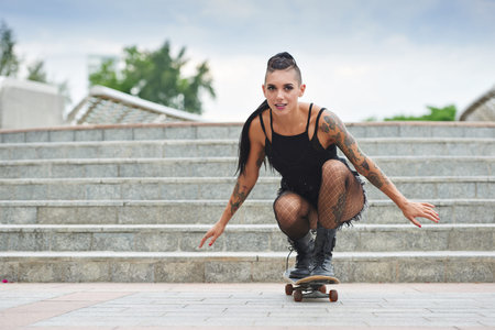Stylish young woman with tattoo skateboarding in the city