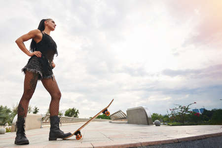 Stylish girl in shorts and fishnet stockings standing with one boot on the edge of skateboardの写真素材